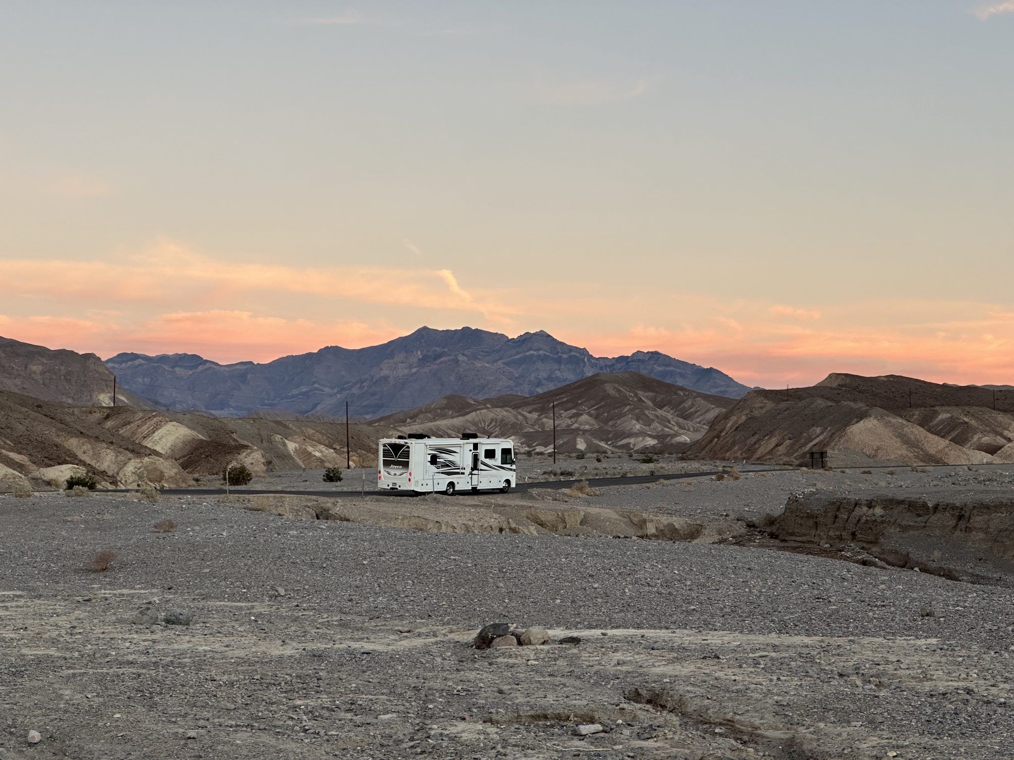 A motorhome on a lonely road through the Death Valley badlands, mountains glowing pink and orange at sunset